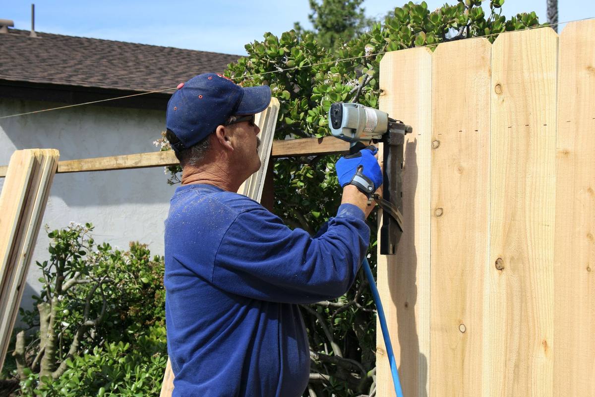 Man in blue shirt and cap uses a nail gun to build a wooden fence outdoors.