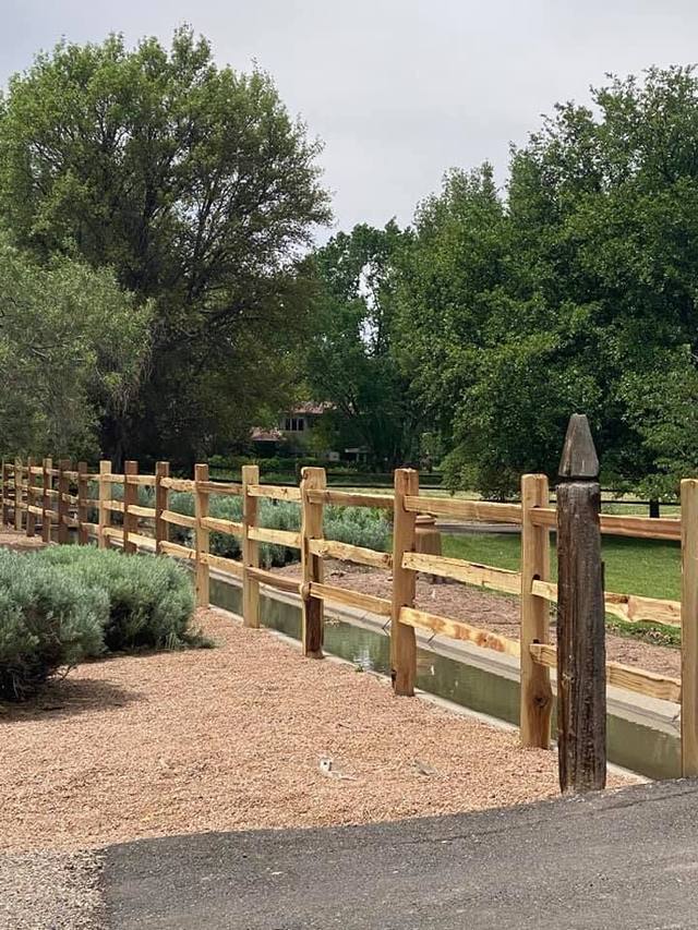 Wooden split-rail fence alongside a gravel path and green shrubbery, with trees in the background under a cloudy sky.