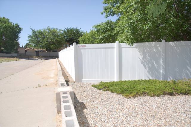 White vinyl fence along a concrete sidewalk next to a gravel bed with green bushes and trees in the background.