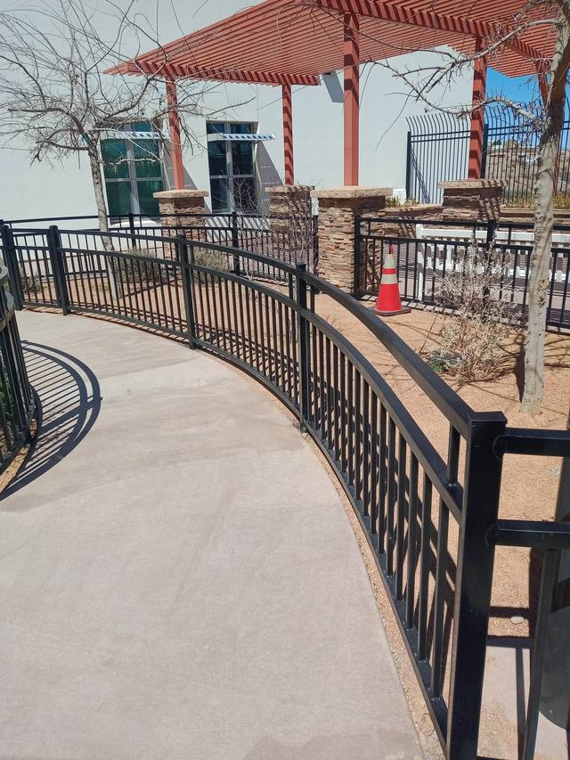 A curved concrete pathway with a black metal fence, leading towards a shaded structure with a red roof.