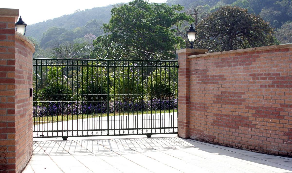 Brick entrance with black metal gate, topped with lamps, leading to a garden with mountain backdrop.