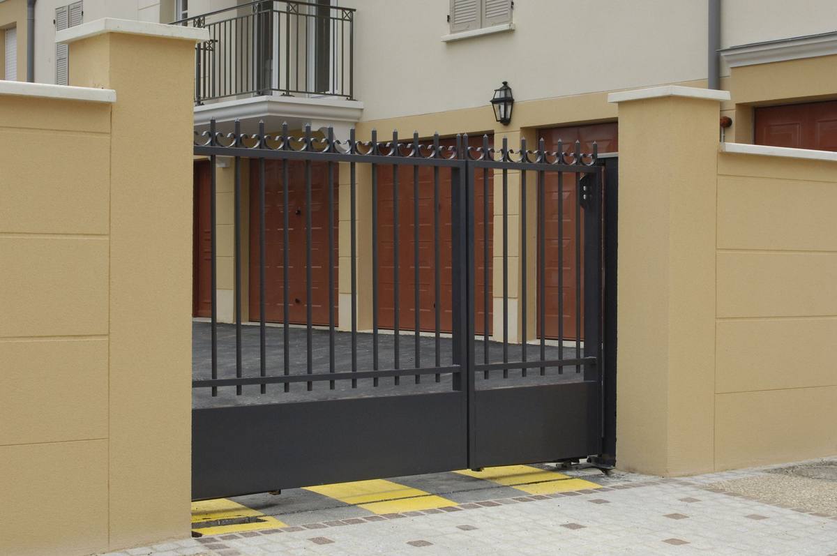 Black metal gate at the entrance to a building. Tan pillars, brown doors in the background. Pavement in front.
